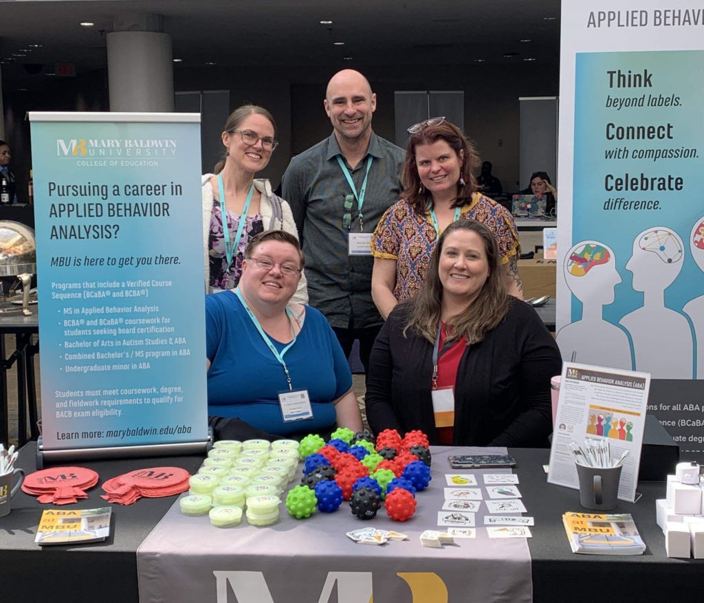 Five professors posing at a fair with MBU Applied Behavior Analysis materials displayed