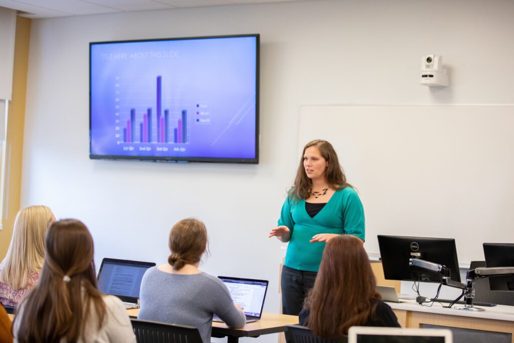 A professor stands and lectures at the front of a class while a presentation on the screen behind them.