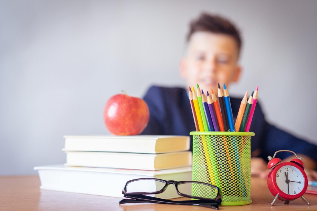 A boy sits behind a series of schools supplies including books, glasses, colored pencils, a clock, and an apple.