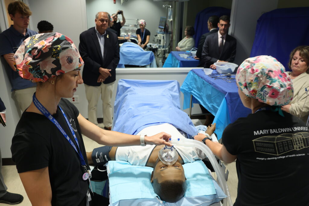 A crowded operation room with instructors, students, and guests surrounding an operating table with a medical simulation Mannequin.