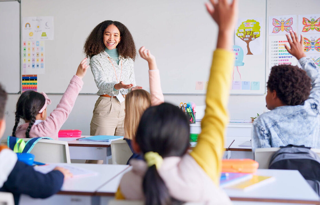 Teacher at front of the classroom with kids raising their hands