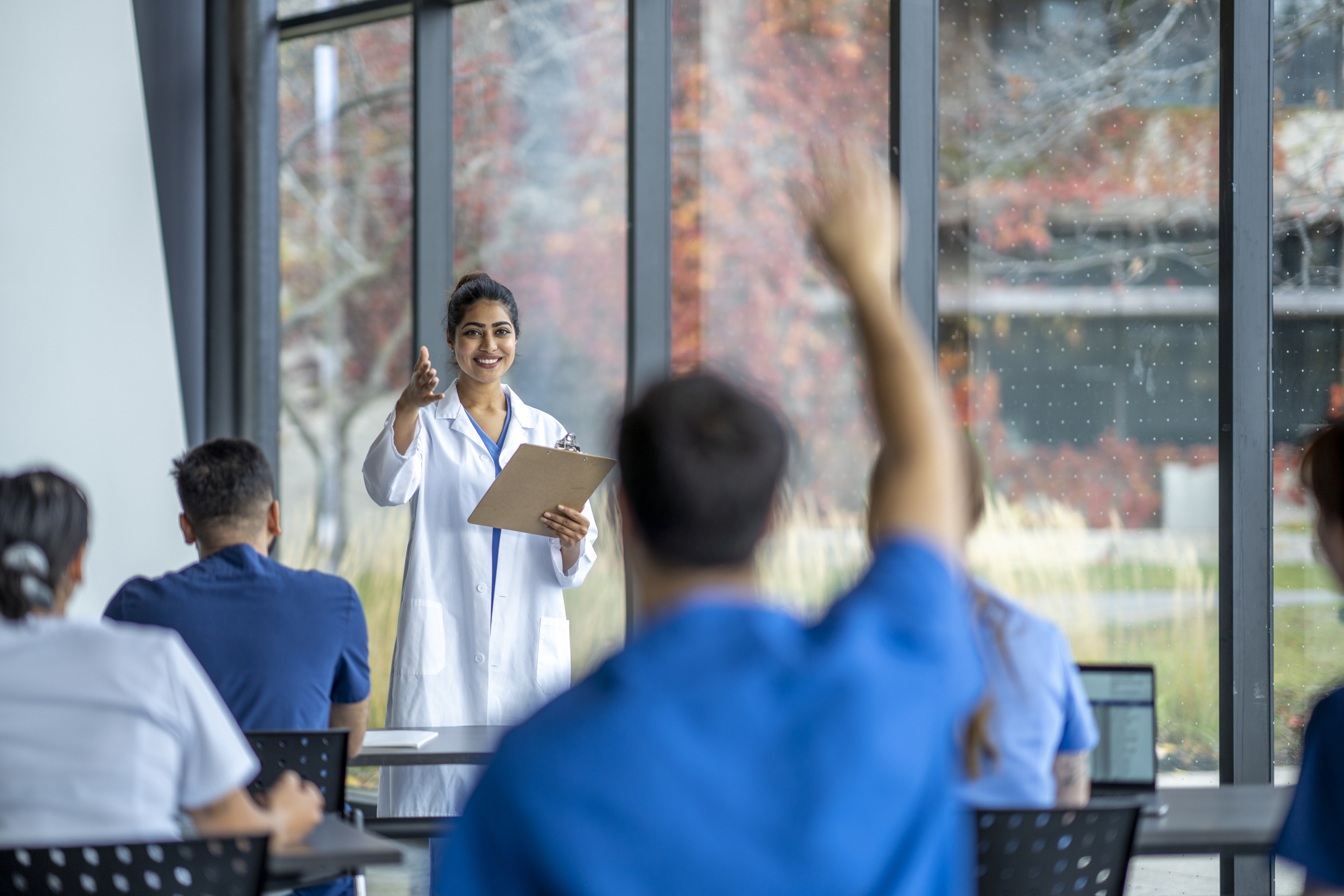 A female doctor and professor stands at the front of a classroom as she teaches a small group of medical students.  She is wearing a white lab coat and the students   are seated in front of her in scrubs.