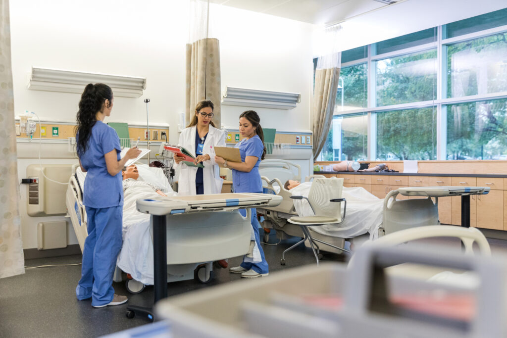 The female hospital trainer stands with the two interns in the medical classroom and repeats instructions so they can confirm their notes are correct.