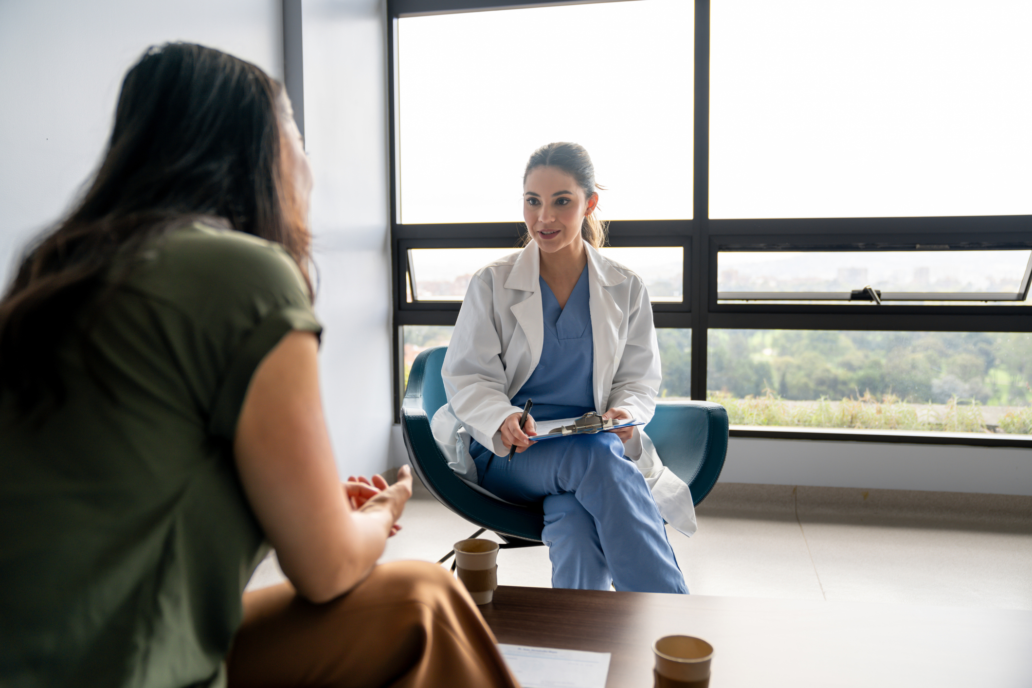 Latin American female doctor talking to a patient in a consultation at the hospital while taking notes on a clipboard