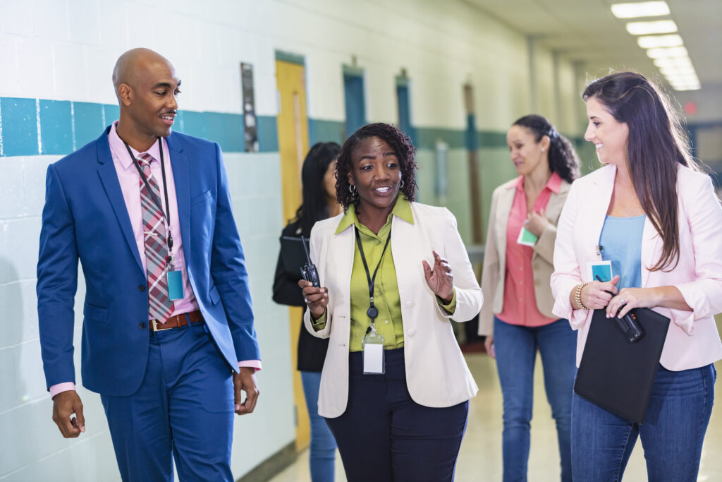 A multiracial group of five teachers or school administrators conversing as they walk through a school corridor. The focus is on the three in the foreground. The African-American woman in the middle, in her 50s, is talking while her coworkers look at her and listen.