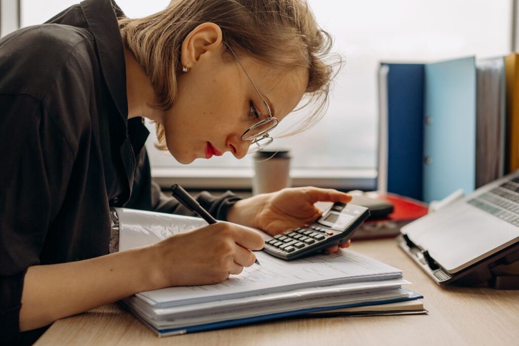 woman with glasses looking at calculator and writing in notebook