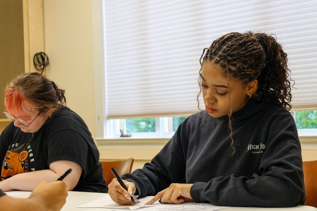 A student writes with a calligraphic pen on a sheet of paper, copying from a nearby hebrew character guide